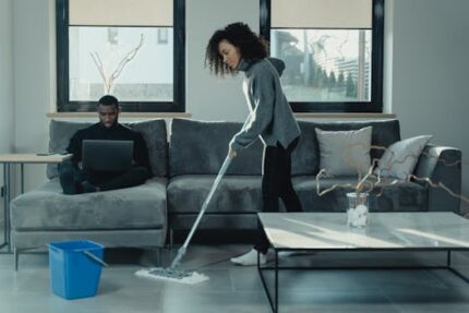 Man working on laptop while woman mops floor in stylish living room.